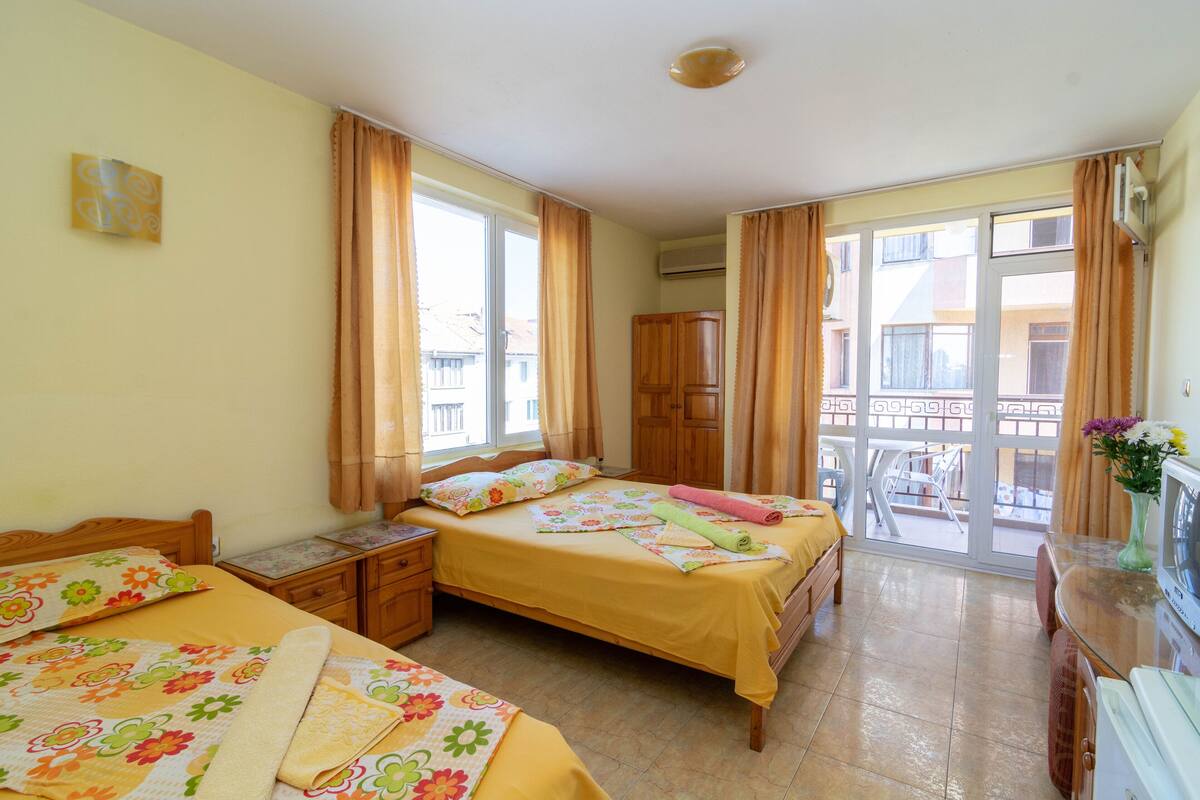 A bedroom with two beds, a wooden wardrobe, a TV, and a balcony with a view of buildings.