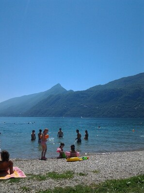 Una spiaggia con persone che nuotano e giocano, un litorale di ciottoli e montagne sullo sfondo.