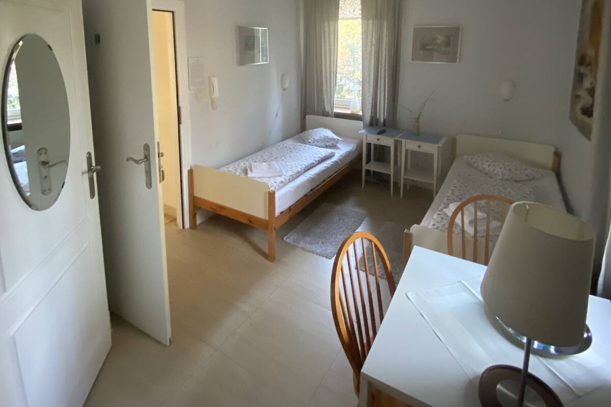 A small, well-lit room with a single bed, a desk, and a chair. There is a door leading to another room, and a window with curtains.