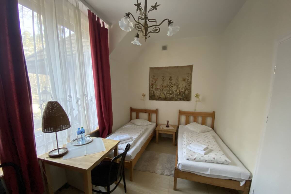 A small, tidy room with two single beds, a desk with a chair, a table with a lamp, and a painting on the wall.