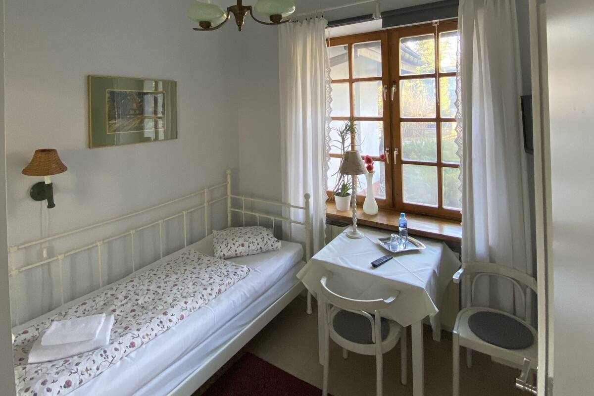 A single-bed room with a small table and two chairs, a window with curtains, a ceiling light, and a wall-mounted lamp.