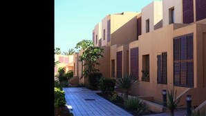A row of beige buildings with dark shutters, a paved walkway, and some greenery.