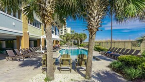 A pool area with lounge chairs and palm trees, surrounded by buildings. - MVG 909 Oceanfront NEW UPDATES (North Myrtle Beach)