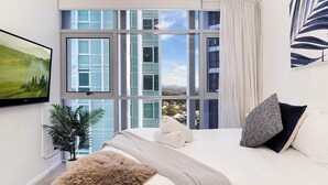A hotel room with a large window, a bed with white bedding, a plant, and a painting on the wall. - River & City Views | Premium Family Apartment (Brisbane City)