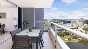 A modern balcony with a glass table and chairs, overlooking a cityscape and a river. - River & City Views | Premium Family Apartment (Brisbane City)