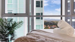 A room with a large window, a plant, and a rolled towel on a bed. - River & City Views | Premium Family Apartment (Brisbane City)