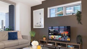 A modern living room with a beige sofa, a glass coffee table, and a flat-screen TV displaying 'Stranger Things'. There are plants and books on shelves, and a framed picture on the wall. - River & City Views | Premium Family Apartment (Brisbane City)