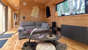 Ein gemütliches Cabin-Interieur mit einem grauen Sofa, einem gewebten Couchtisch und einem großen Fenster mit Blick auf Bäume.