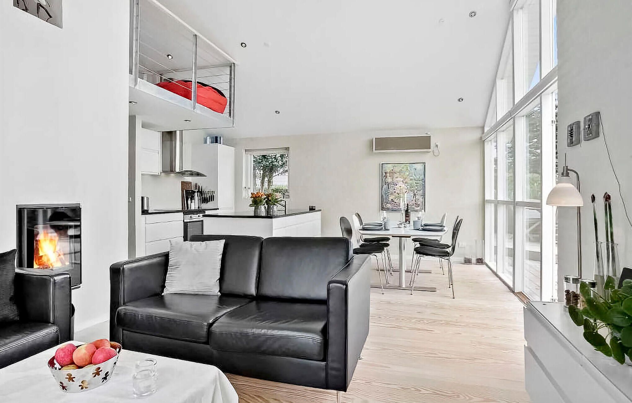 A modern living room with a black leather sofa, a dining area with a table and chairs, and a kitchen with white cabinets.