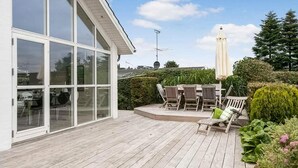En träaltan med utemöbler, en vit byggnad med stora fönster och en satellantenn.