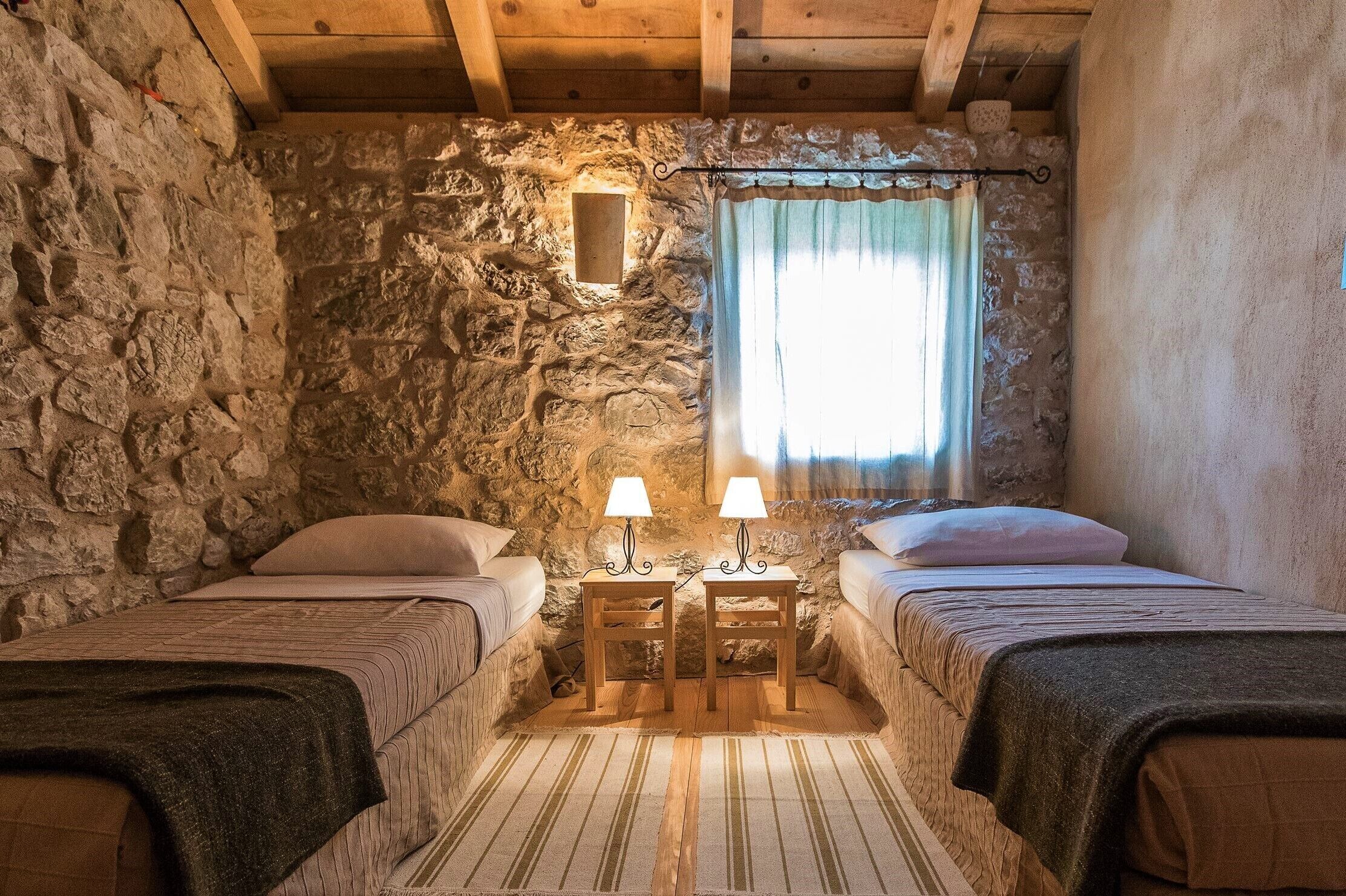 A rustic room with two beds, stone walls, wooden beams, a small window with curtains, and a wooden nightstand with lamps. - Authentic stone built villetta with a magnificent view (Grižane)