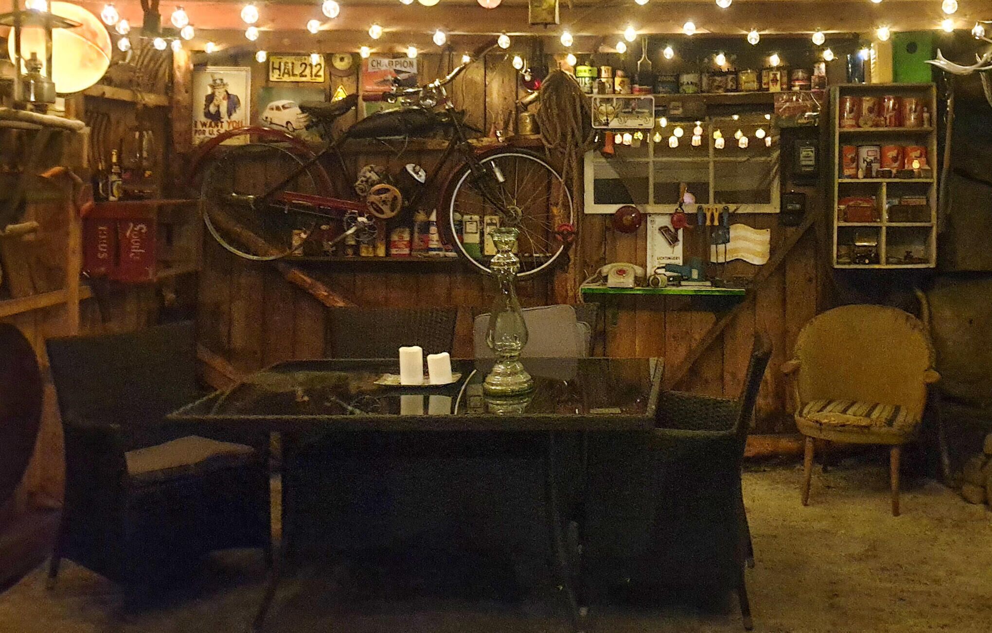 A cozy bar area with a bicycle mounted on the wall, a glass table with a candle, and shelves stocked with various items.