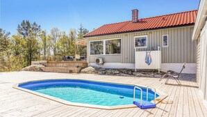 Een zwembad met een houten terras, een overdekte patio met zitplaatsen, en een huis met een rode pannendak.