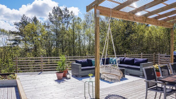 Een houten terras met buitenmeubilair, zoals een bank en stoelen, onder een pergola met een metalen dak.