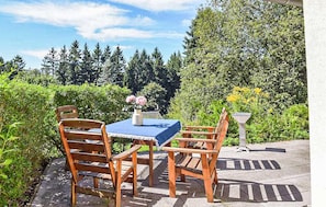 Une table en bois placée à l’extérieur, avec des chaises, une nappe bleue et un vase rempli de fleurs.