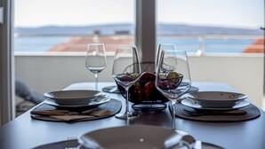 Une table à manger dressée avec des assiettes, des couverts et des verres à vin, agrémentée d’un panier de fruits et offrant, en arrière-plan, une vue sur la mer et les montagnes.