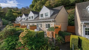 A row of houses with stone and wood exteriors, neatly arranged on a hillside with well-maintained gardens and hedges. - Conveniently located to explore all that Cornwall and Devon have to offer (SALTASH)