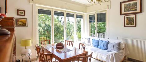 A dining area with a wooden table and chairs, a chandelier, and a view of greenery through large windows.