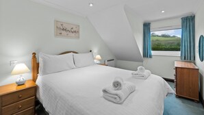 Een slaapkamer met een bed, nachtkastje, kapstok, raam met gordijnen en een aan de muur hangend schilderij.