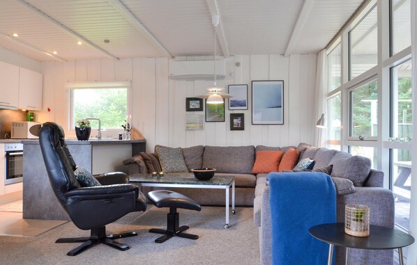 Un salon moderne avec un canapé gris, un fauteuil en cuir noir et une table basse. On aperçoit, au fond, un coin cuisine avec un four à micro-ondes et un évier.