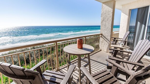 A balcony with a wooden table and chairs, overlooking a beach with waves.