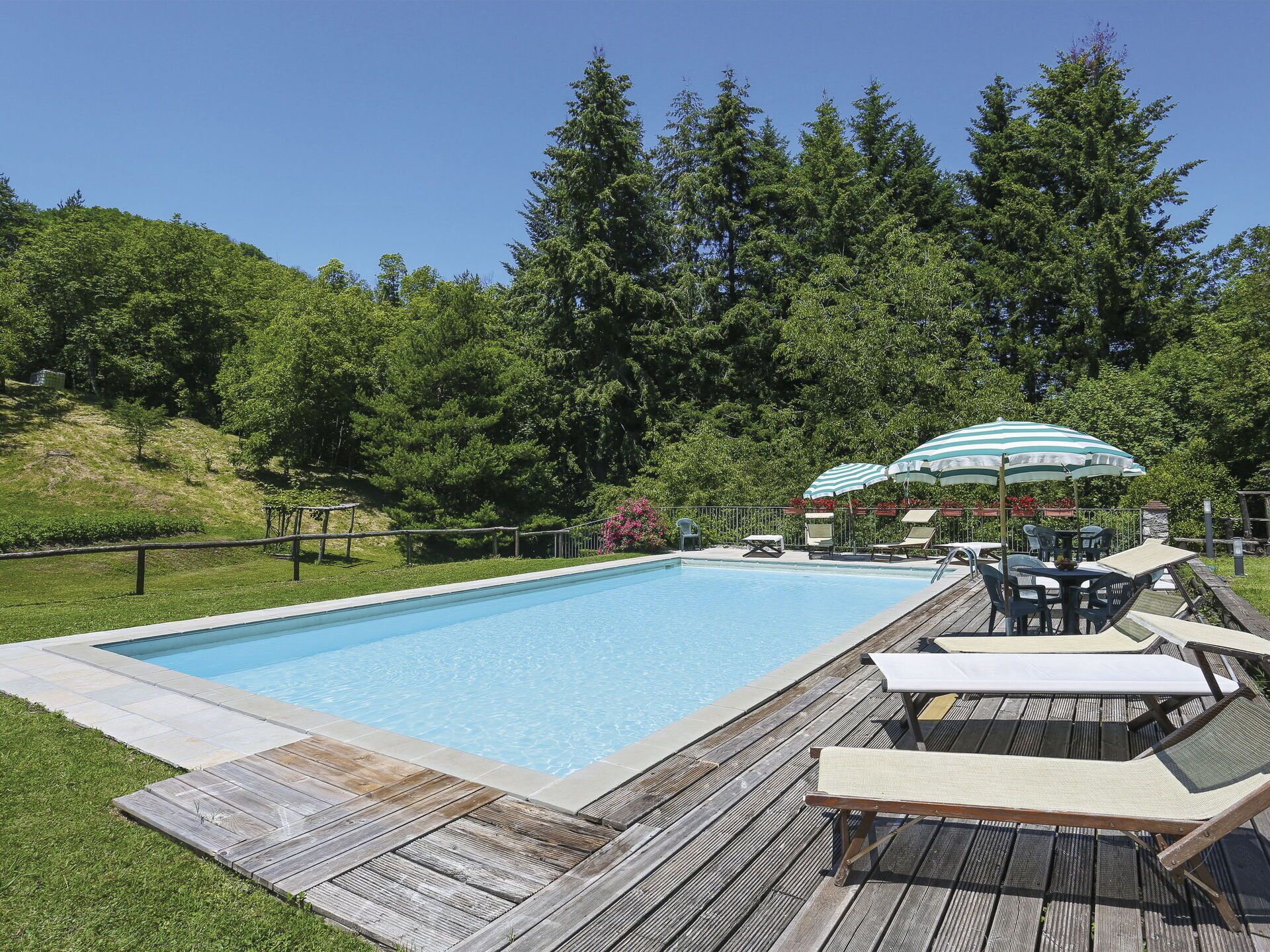 Ein rechteckiger Pool mit umlaufender Holzterrasse, ausgestattet mit Liegestühlen und Sonnenschirmen.