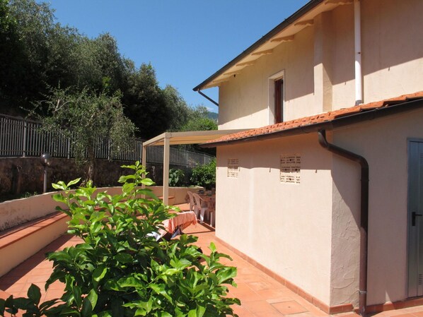 Ein Gebäude mit einem Ziegeldach und einer überdachten Terrasse. Im Vordergrund befinden sich Pflanzen, und im Hintergrund ist ein Zaun zu sehen.