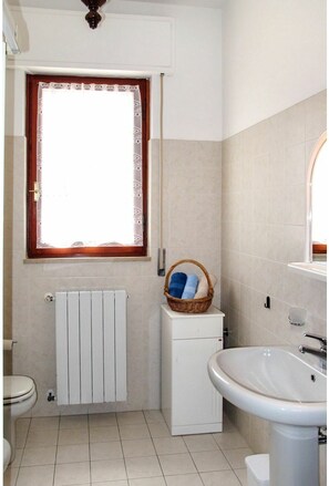 Ein Badezimmer mit Toilette, einem weißen Heizkörper, einem weißen Schrank, einem Korbwäscheschalen mit Handtüchern und einem Waschbecken.