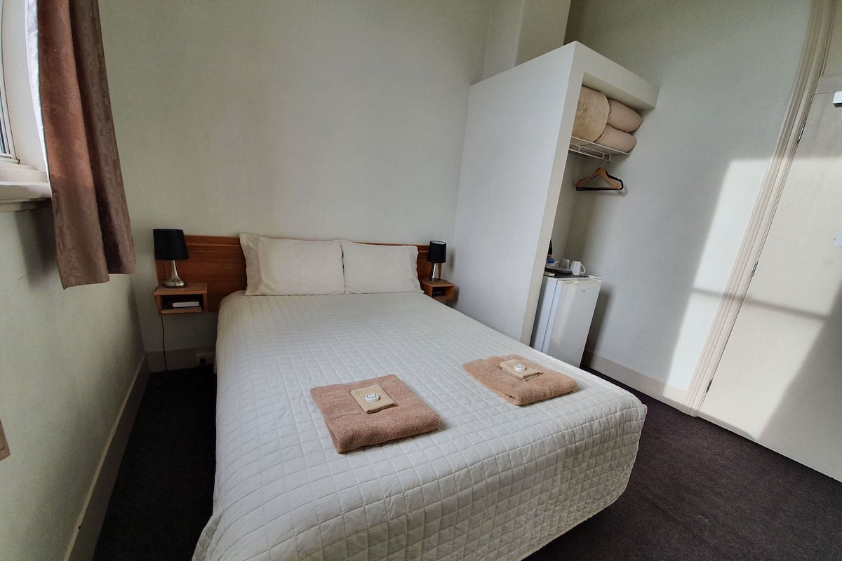 A neatly made bed with a quilted cover, a bedside table with a lamp, a wardrobe, and a small shelf with a cup and a book.