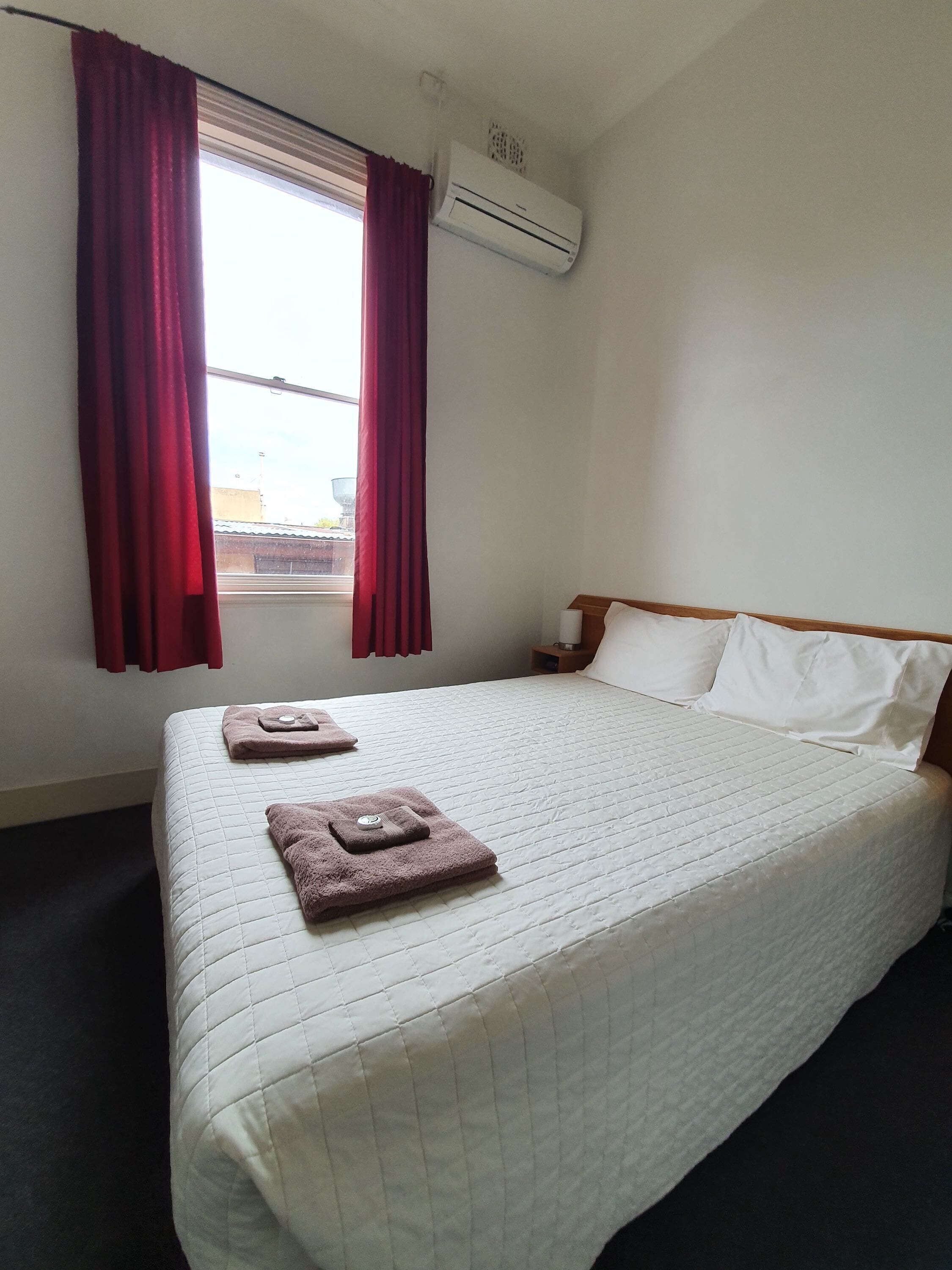 A hotel room with a bed, red curtains, an air conditioning unit, and a window.