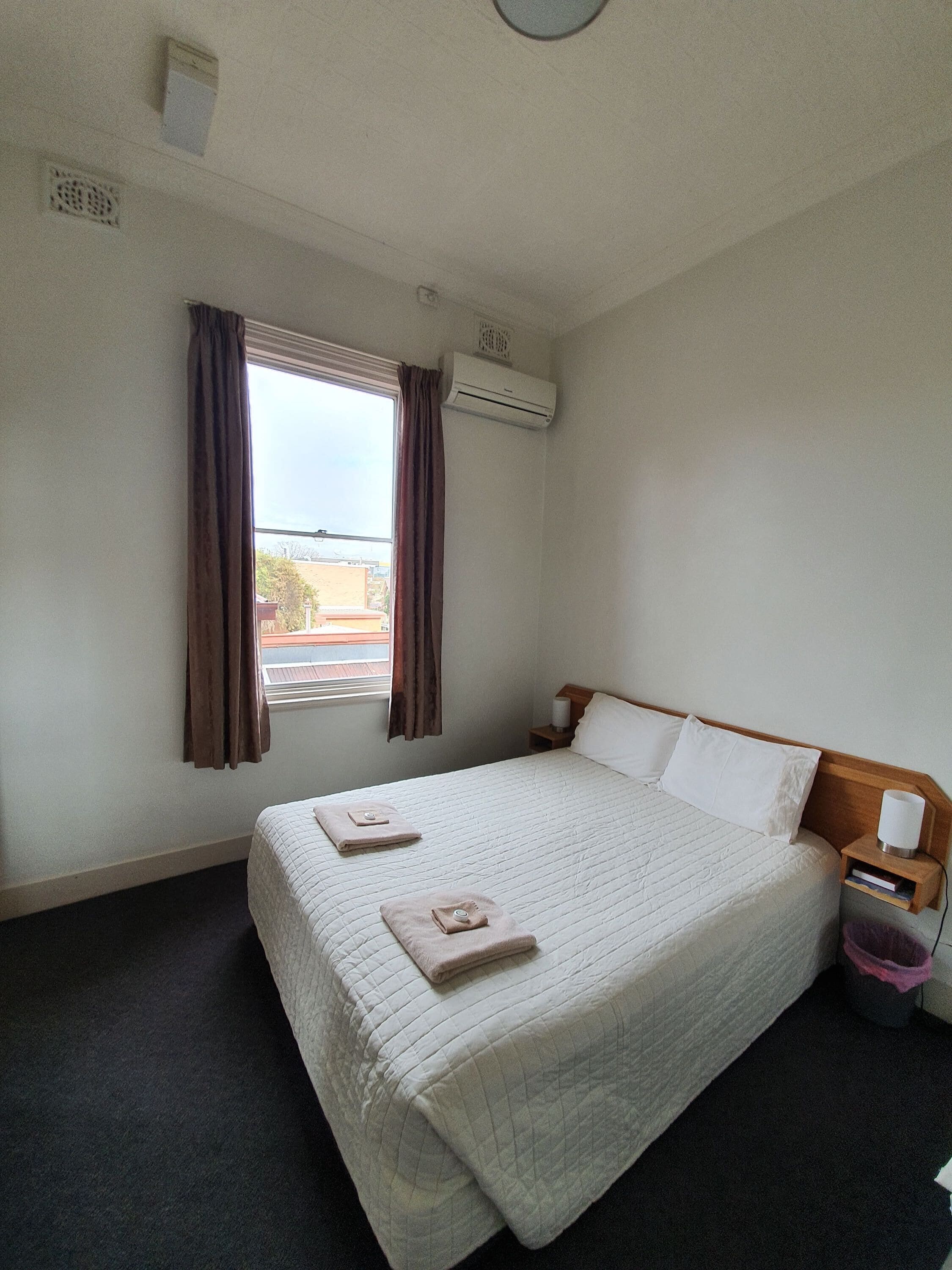 Una habitación de hotel con una cama grande, una ventana con cortinas, un equipo de aire acondicionado y una mesita de noche con lámpara.
