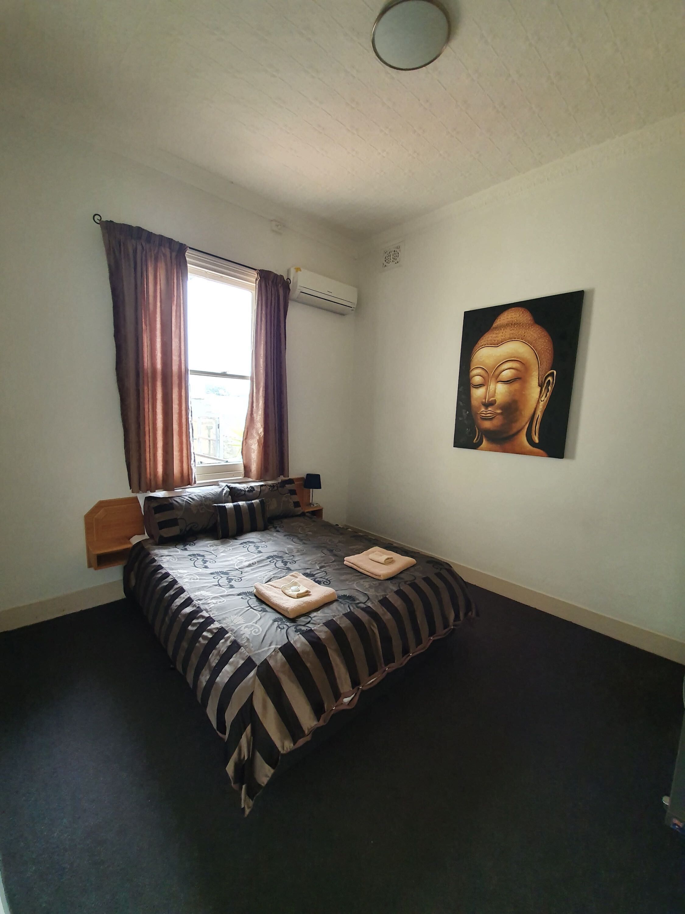 Un dormitorio con cama, ventana con cortinas, equipo de aire acondicionado, mesita de noche, lámpara, pintura de Buda colgada en la pared y un pequeño estante de madera.