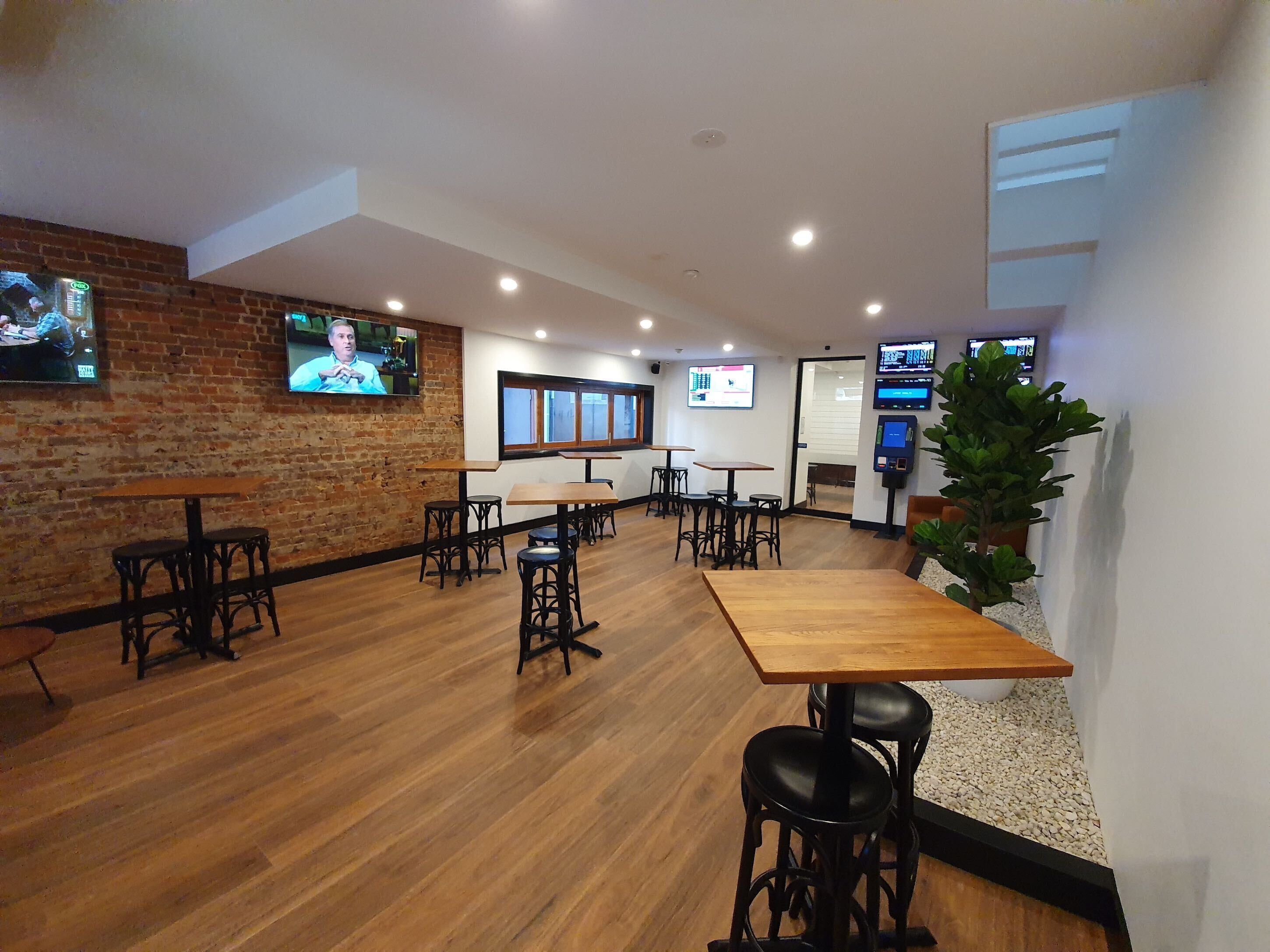Un espace bar moderne avec des tables et des tabourets en bois, des murs en briques et plusieurs écrans diffusant du contenu.