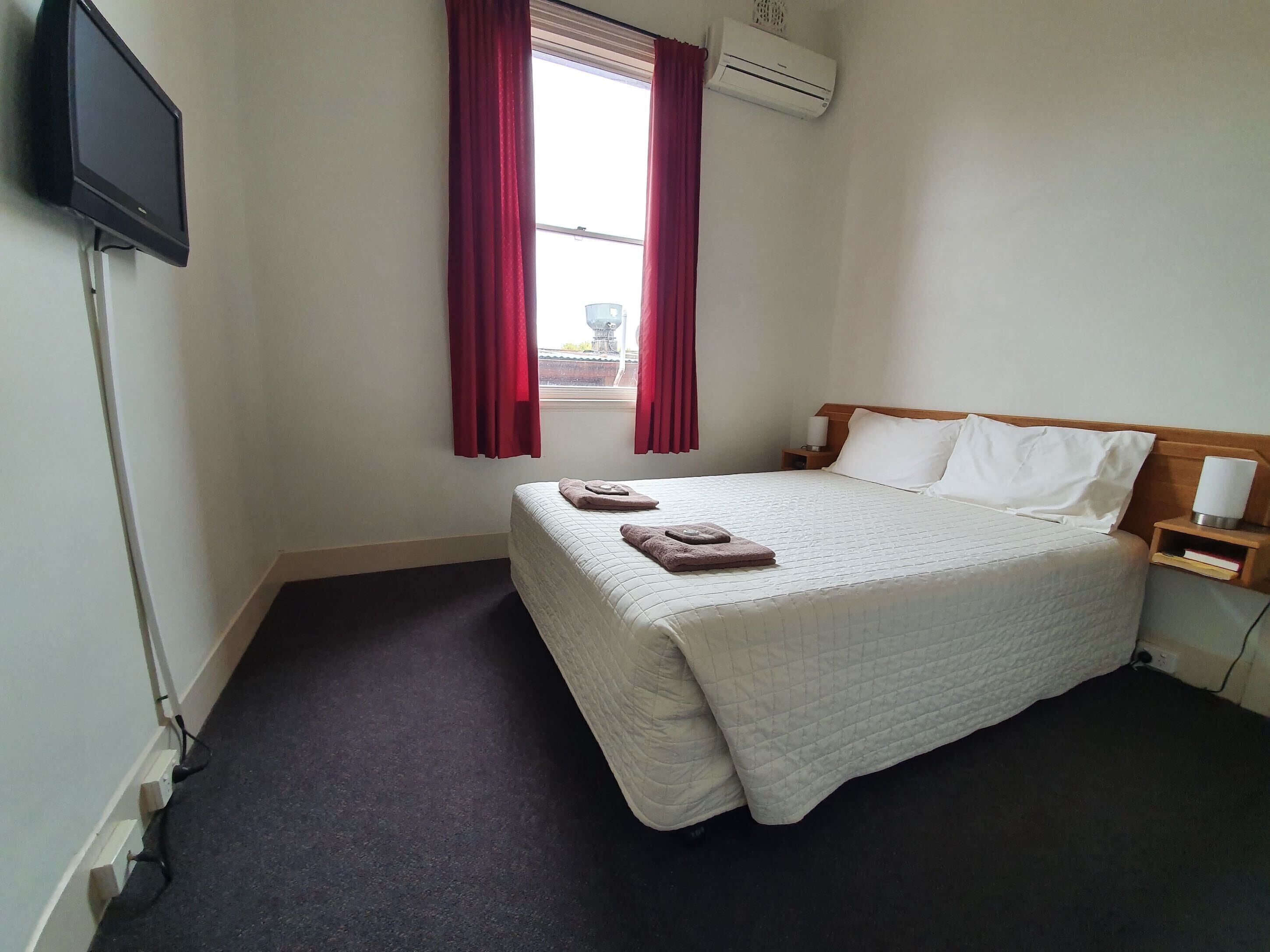A hotel room with a bed, a bedside table with a lamp, a TV mounted on the wall, and an air conditioning unit.