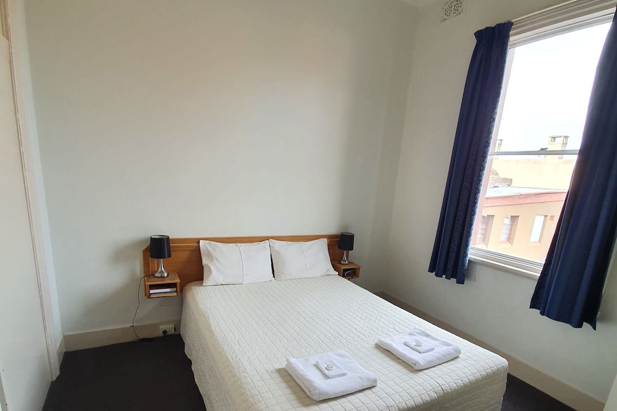 A neatly made bed with white linens and towels, a bedside table with a lamp, and a window with curtains.