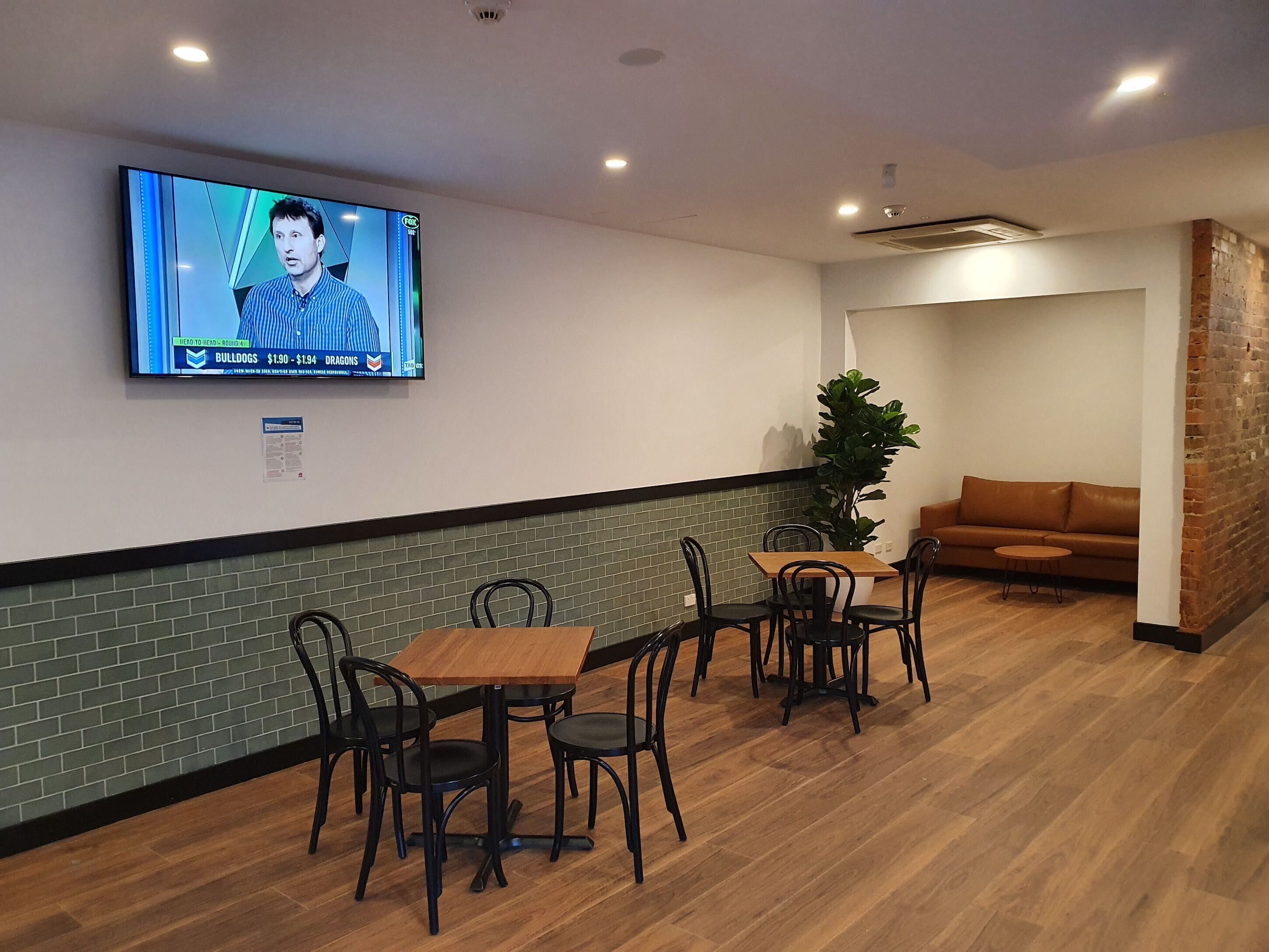 A modern lounge area with a television mounted on the wall displaying a news program, a brick accent wall, a wooden floor, and a brown leather sofa.