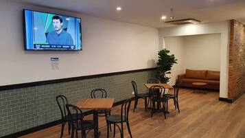 A modern lounge area with a television mounted on the wall displaying a news program, a brick accent wall, a wooden floor, and a brown leather sofa.