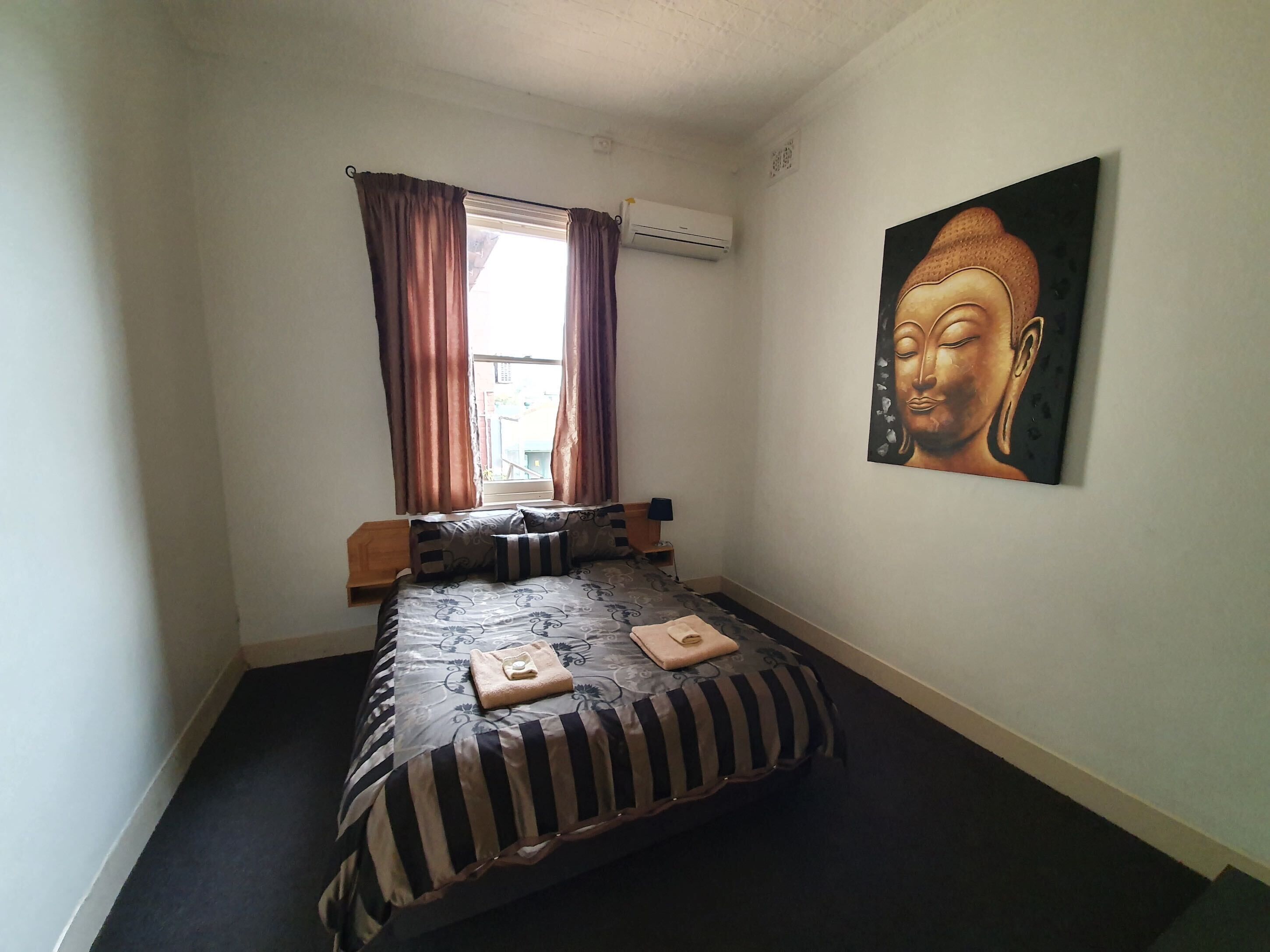 A small, neatly arranged bedroom with a bed, a window with curtains, an air conditioning unit, and a Buddha painting on the wall.