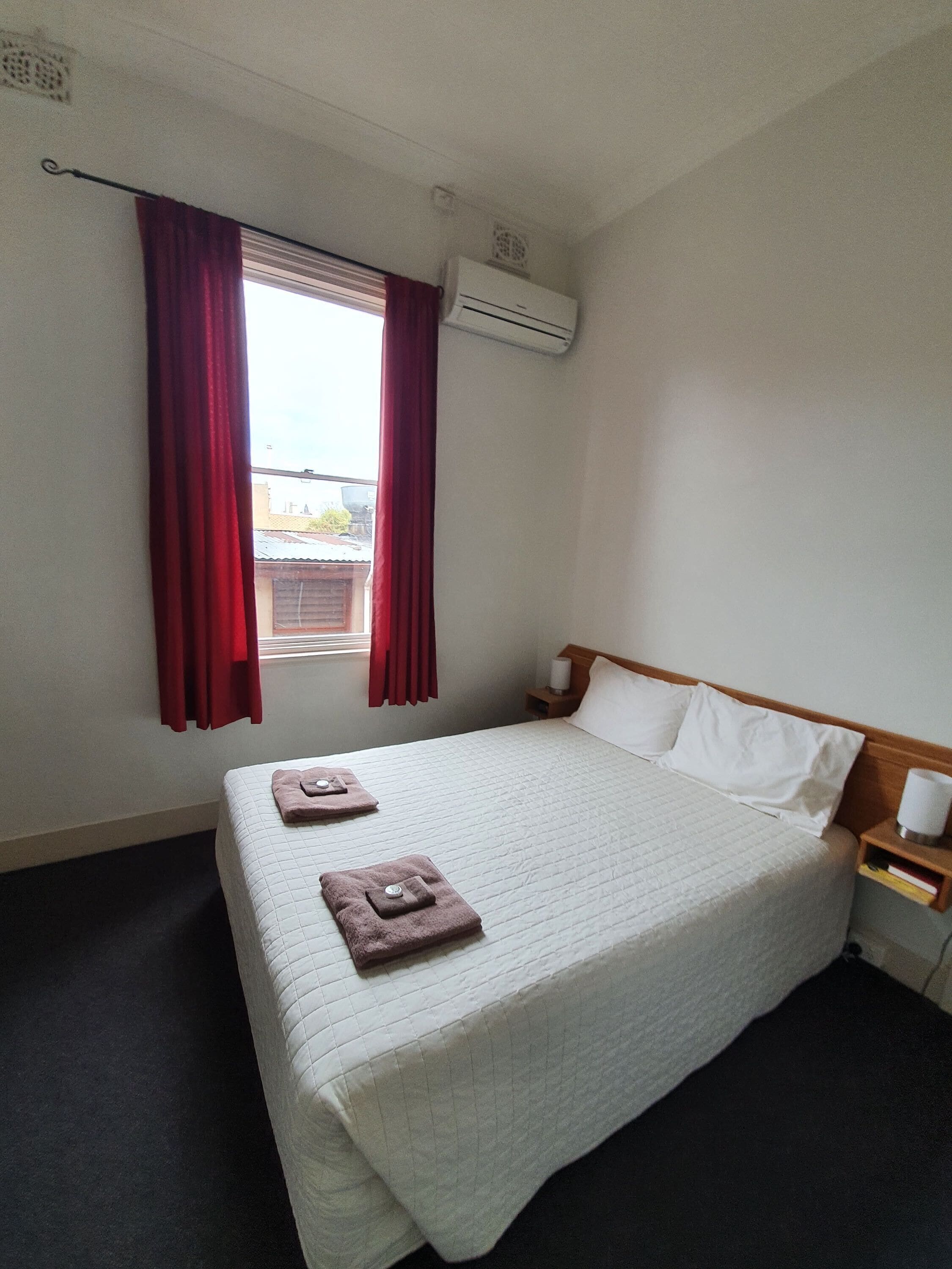 A hotel room with a bed, a window with red curtains, an air conditioning unit, and a bedside table with a lamp and a book.