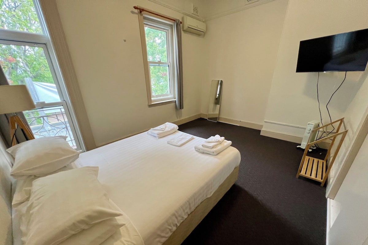 A hotel room with a bed, pillows, a window, an air conditioning unit, a television mounted on the wall, and a small wooden chair.