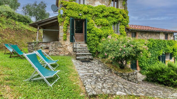 A stone house with a sloped roof, ivy-covered walls, and a satellite dish. There are two green lounge chairs in the front yard. - Lovely apartment for 4 guests with WIFI, pool, TV, patio and pets allowed (Pescia)