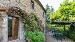 A stone building with a wooden door and a number '4' sign. There is a garden with pink flowers and green bushes. A table and chairs are under a wooden pergola. - Lovely apartment for 4 guests with WIFI, pool, TV, patio and pets allowed (Pescia)