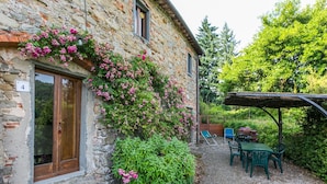 A stone building with a wooden door and a number '4' sign. There is a garden with pink flowers and green bushes. A table and chairs are under a wooden pergola. - Lovely apartment for 7 guests with pool, WIFI, TV, patio and pets allowed (Pescia)