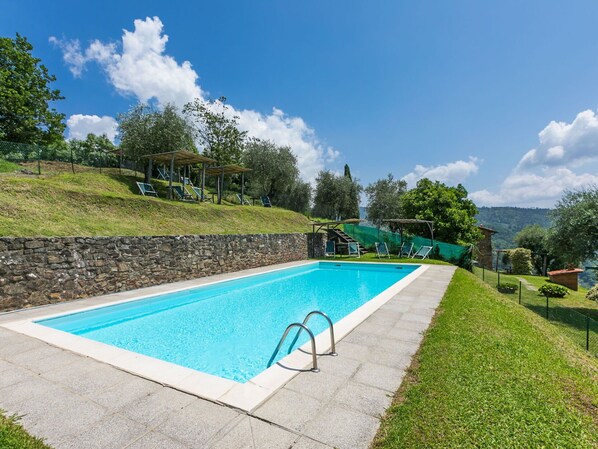 Ein rechteckiger Pool mit einer Steinmauer an einer Seite und einer Rasenfläche mit Liegestühlen und einer Pergola auf der anderen Seite.