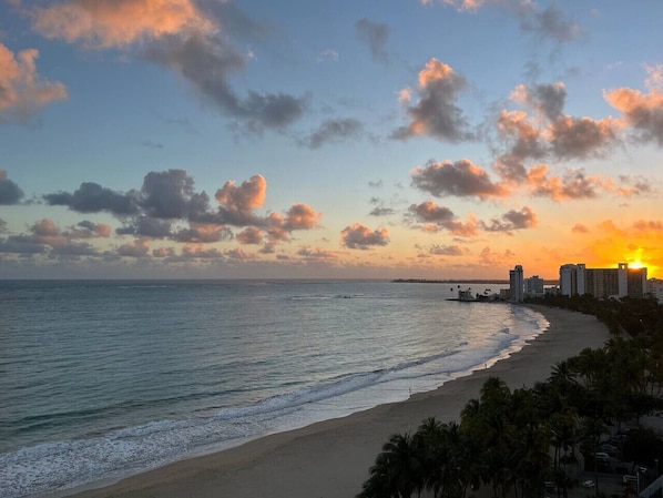 A beach with a sunset, ocean, and buildings in the background.