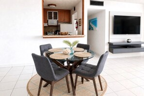 A modern living room with a glass dining table, grey upholstered chairs, a flat-screen TV, and a kitchen visible through an open doorway.