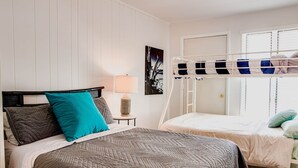 A bedroom with two beds, one with a bunk bed above it, a bedside table with a lamp, and a framed picture on the wall.