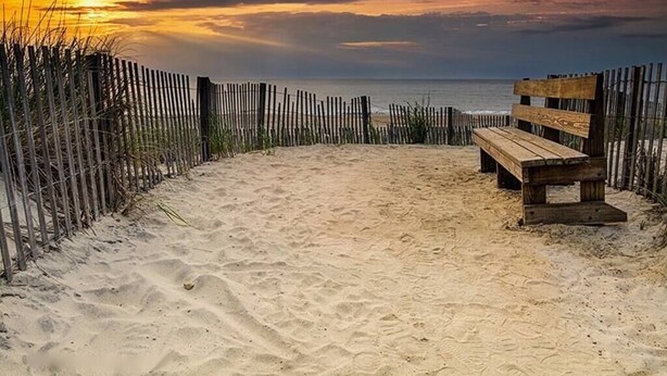 A wooden bench on a sandy beach with a wooden fence and a view of the ocean.