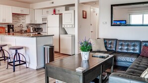 A modern kitchen with white cabinets, a central island with stools, a microwave, and a refrigerator. There is a dark wooden table with a plant and a remote control. A black leather sofa is visible in the background.