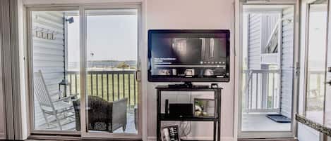 Uma sala de estar com televisão sobre um móvel, uma porta de vidro deslizante para uma varanda e uma pequena mesa.